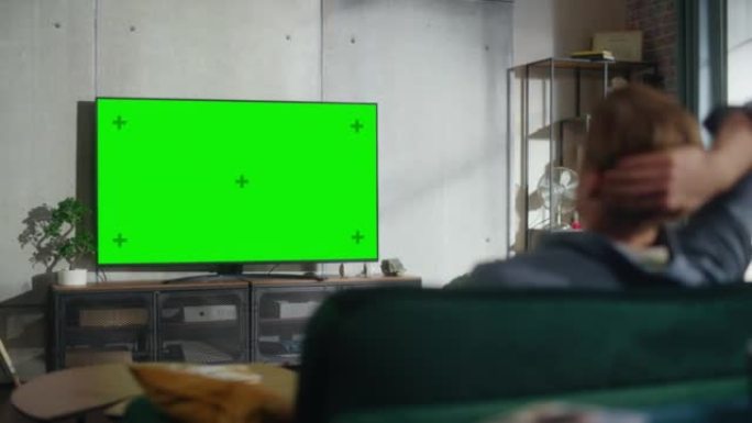 年轻英俊的男人坐在沙发上，看着电视，水平的绿屏模拟。这是阁楼公寓家里客厅的白天。