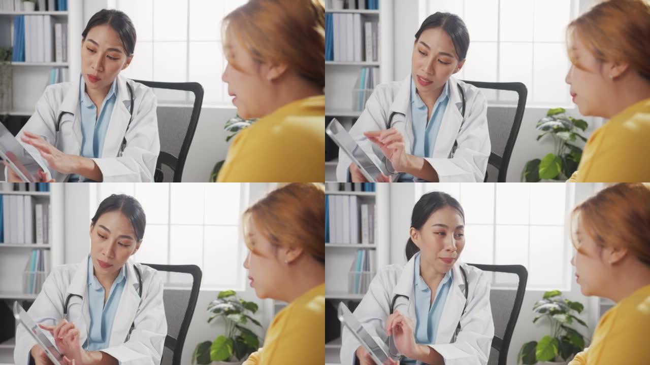 年轻的亚洲女性医生在白色医疗制服使用平板电脑正在传递伟大的新闻谈话讨论结果或症状与女孩患者坐在办公桌