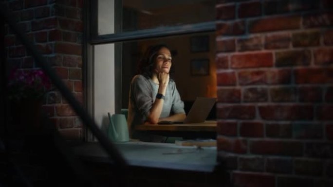 微笑的西班牙女人在温暖舒适的晚上在家使用笔记本电脑。专业自由职业者在家远程工作。从外面拍摄到公寓的窗