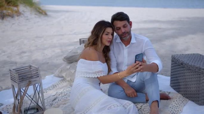 美丽的成年夫妇在海滩上躺在野餐毯上放松，一边看智能手机上的社交媒体，一边聊天