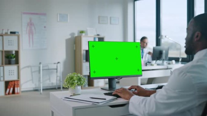 非裔美国医生正在健康诊所使用带有绿屏模拟显示的计算机。穿着白大褂的助理正在医院办公室的桌子后面阅读病