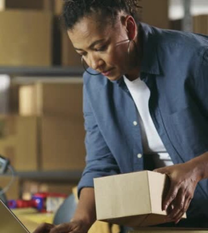 垂直屏幕: 库存经理准备一个小纸板包裹进行邮资。多种族的非裔美国女性小企业主在仓库的笔记本电脑上工作