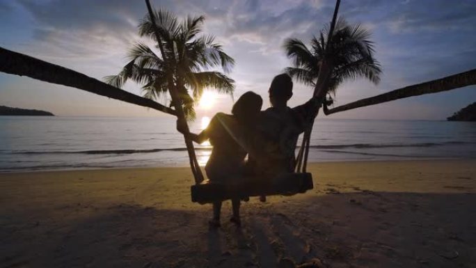 海滩上可爱的夫妇的剪影。年轻的潮人男人和女人坐在一起在秋千上拥抱和亲吻，同时在日落时一起在海边享受暑
