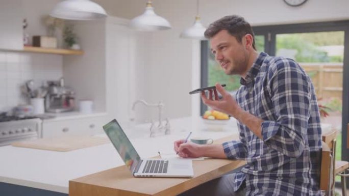 男子在家工作，在厨房柜台上使用笔记本电脑与手机上的麦克风通话