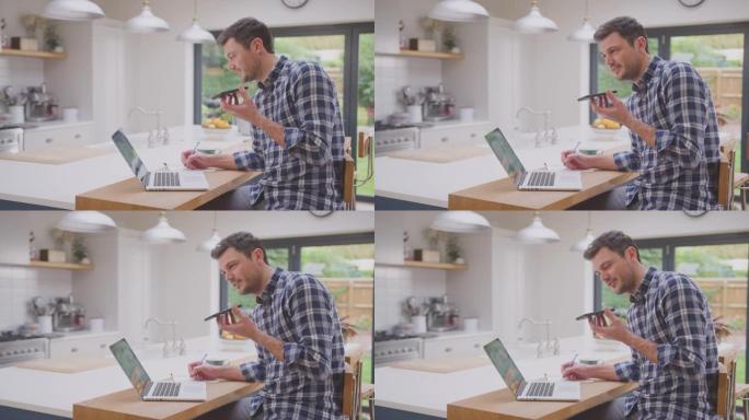 男子在家工作，在厨房柜台上使用笔记本电脑与手机上的麦克风通话