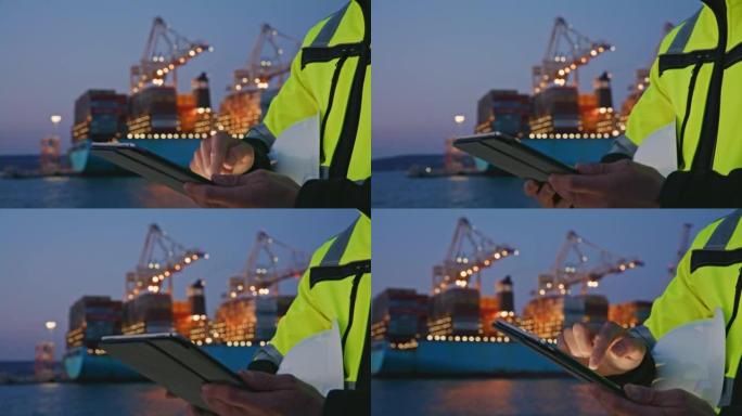 特写工程师，戴着警告背心和头盔的男子在数字平板电脑上打字，晚上站在一艘大型集装箱船前的港口