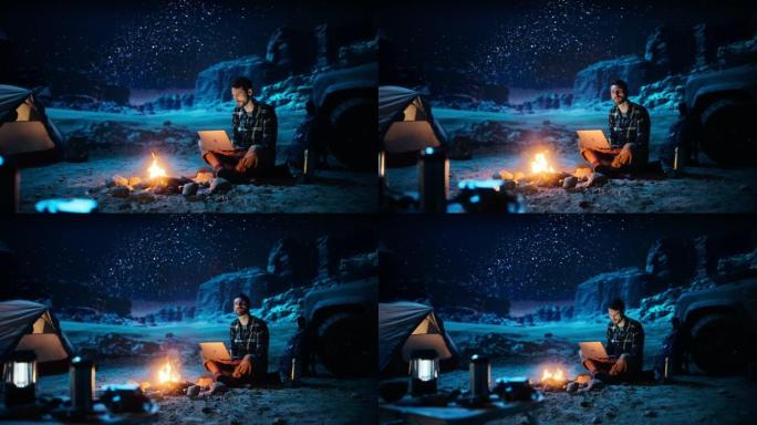 峡谷夜间帐篷露营: 男性旅行者使用坐在篝火旁的笔记本电脑。从事数字远程工作，电子购物，电子商务，使用