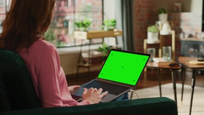 年轻的成年女性在带绿屏模拟显示的笔记本电脑上的家庭阁楼公寓工作。使用触控板和浏览互联网的创意女性。特