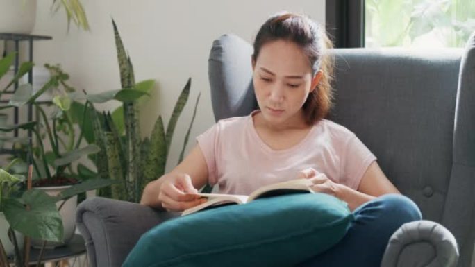 年轻的亚洲大学女性，随意坐在椅子上，在家里的客厅里阅读最喜欢的书课文学习。