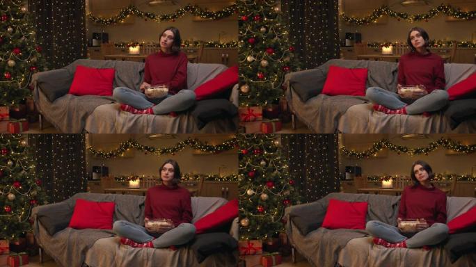 一个穿着红色毛衣和牛仔裤的棕色头发的女人正拿着一碗爆米花看电视剧，担心电影中的人物，坐在装饰有圣诞树