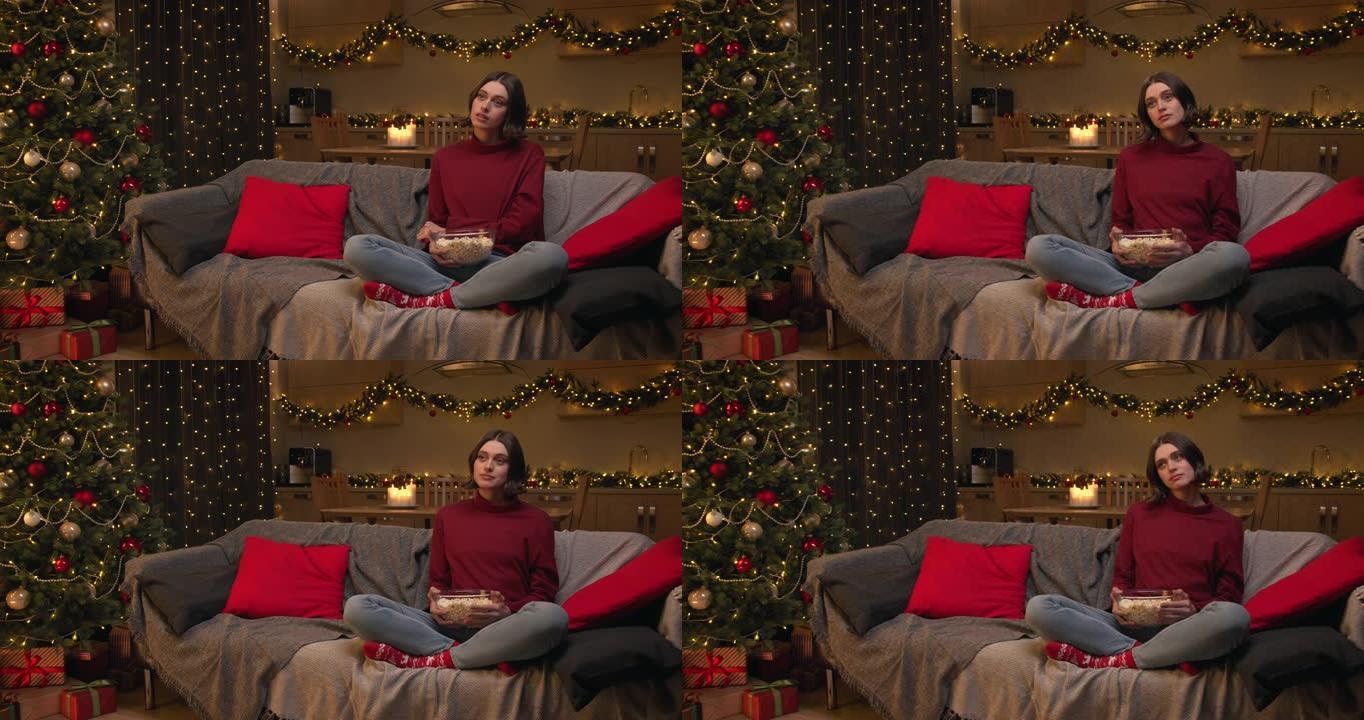 一个穿着红色毛衣和牛仔裤的棕色头发的女人正拿着一碗爆米花看电视剧，担心电影中的人物，坐在装饰有圣诞树