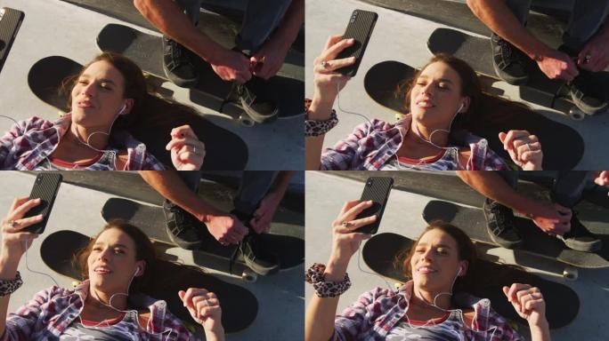 快乐的高加索女人在滑板场使用智能手机和男人系鞋带