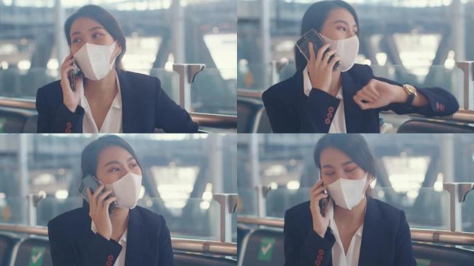 聪明的亚洲商务女孩穿西装，坐在长凳上，带着手提箱打电话给智能电话，与合作伙伴在机场等待航班。