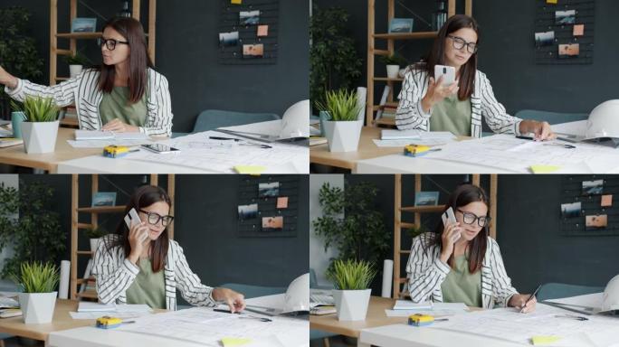 年轻的女建筑师使用蓝图做笔记，然后在办公室用手机通话