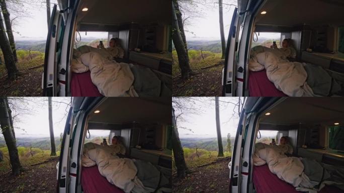 金发碧眼的女人放松下来，躺在面包车内的床垫上，用智能手机，腿上的毯子，广角拍摄，车门打开，摄像头移开