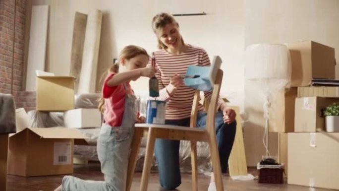 入住和家庭装修: 快乐的母亲画复古家具椅子女儿跑来帮助她。开朗的年轻家庭使公寓充满艺术和色彩。微笑和