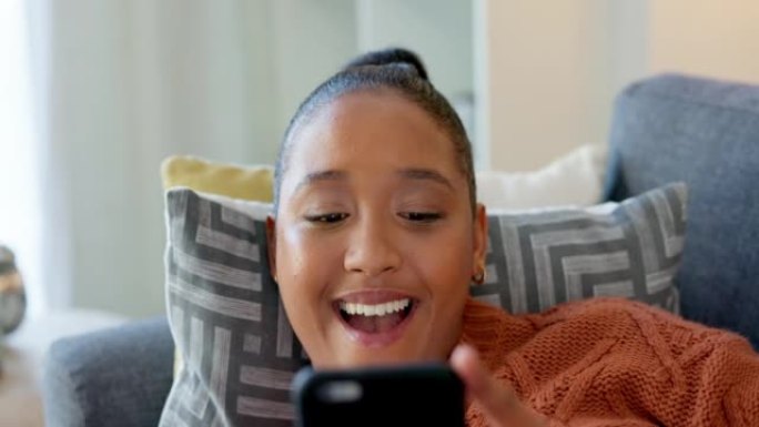 快乐的年轻女子躺在客厅的沙发上使用电话。休闲的非洲女性看起来开朗舒适，喜欢在网上浏览和在家查看社交媒