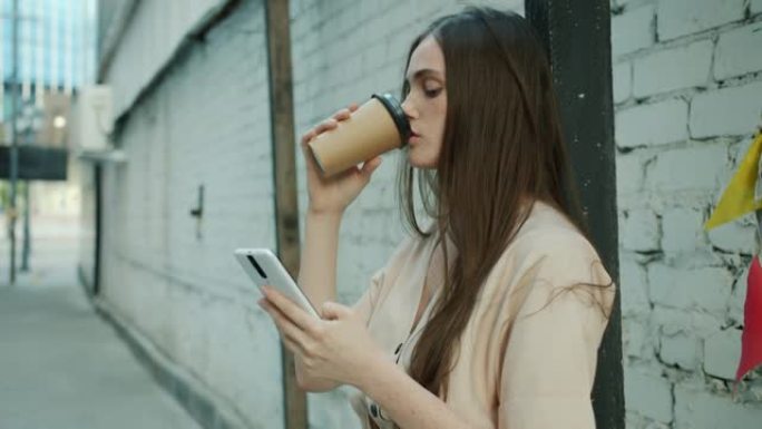 沉思的年轻女子触摸智能手机屏幕，在城市街道户外喝咖啡