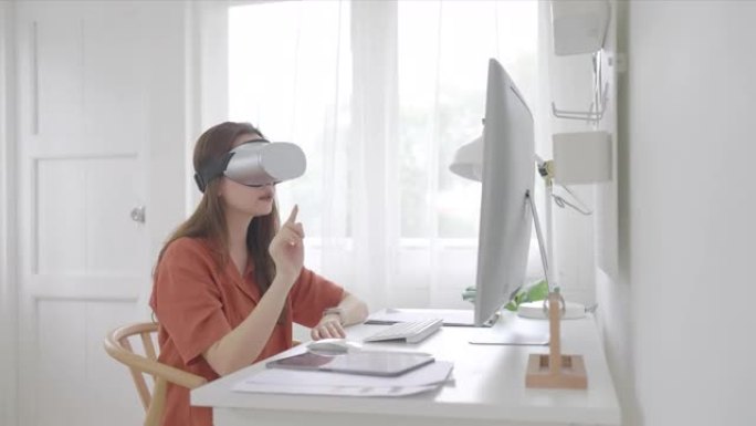 美丽的亚洲女性年龄36岁坐在家庭办公室戴着虚拟现实护目镜，触摸空气在虚拟现实体验和电脑远程工作。成人