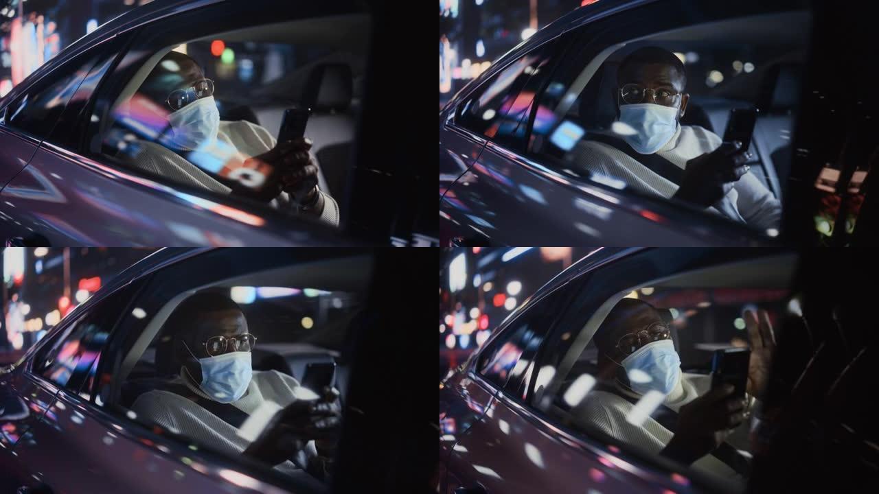 时尚的黑人男子戴着口罩，晚上在出租车后座上通勤回家。英俊的男性在带有工作霓虹灯的城市街道上的转车上，