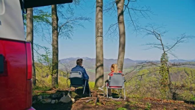 夫妻俩在大自然中露营，既坐在椅子上，又从杯子里喝酒，男人站起来看着风景