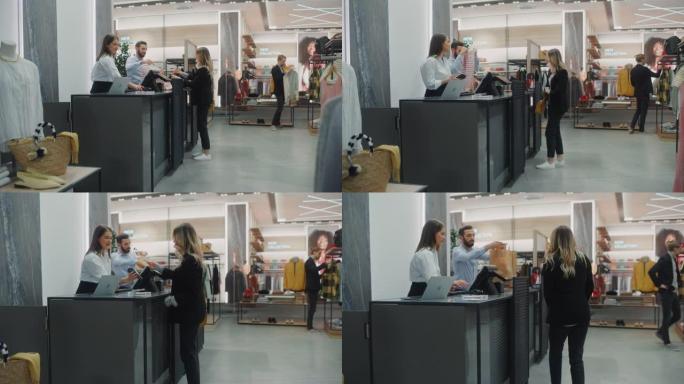 服装店收银台: 美丽的年轻女子从友好的零售助理那里购买衬衫，使用非接触式信用卡付款。拥有设计师品牌的