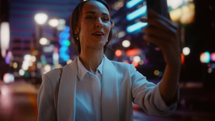 美丽的年轻女子使用智能手机进行视频通话，穿过霓虹灯的夜晚城市街道。使用手机与家人，朋友或商业伙伴聊天