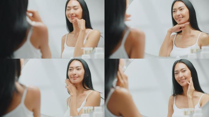 美丽的亚洲女人抚摸着她完美柔软的皮肤和郁郁葱葱的黑发，在浴室的镜子里微笑。健康健康概念，自然美，有机