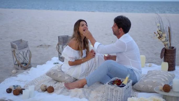 充满爱心的男人在海滩上享受浪漫的野餐时，用水果喂妻子聊天，喝香槟