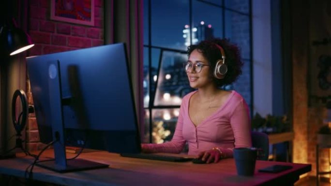 晚上在时尚的阁楼公寓里，戴着耳机的年轻漂亮黑人妇女使用电脑。有创意的女性微笑，在社交媒体上浏览视频。