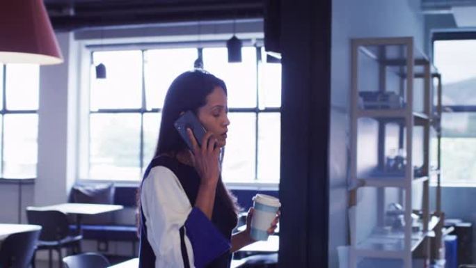 混血女商人在智能手机上聊天，拿着咖啡走在办公室走廊上