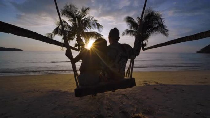海滩上可爱的夫妇的剪影。年轻的潮人男人和女人坐在一起在秋千上拥抱和亲吻，同时在日落时一起在海边享受暑