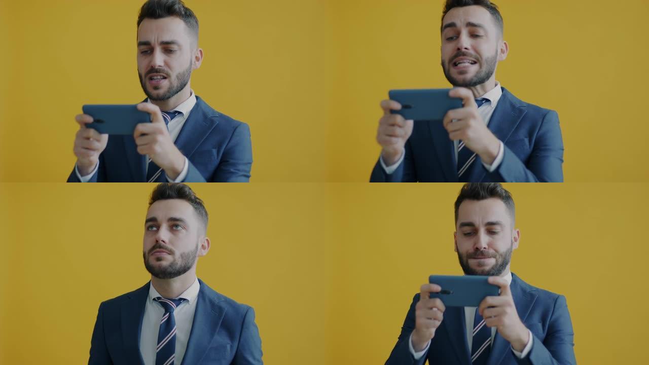 商人用智能手机玩视频游戏的肖像在黄色背景上愤怒地看着屏幕