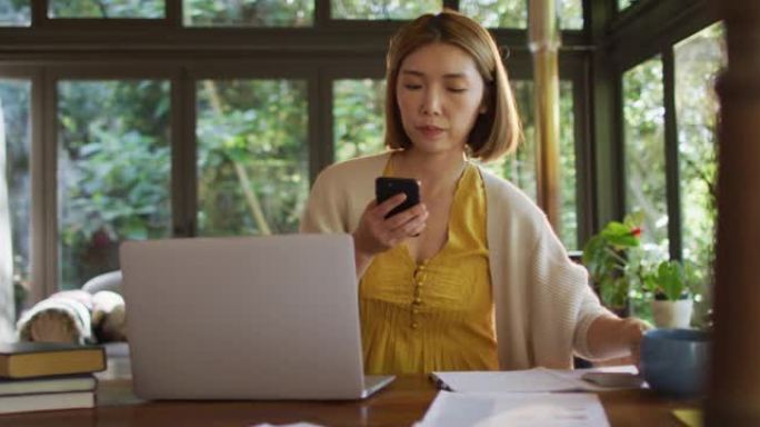 坐在餐桌旁在家工作并使用智能手机的亚洲妇女