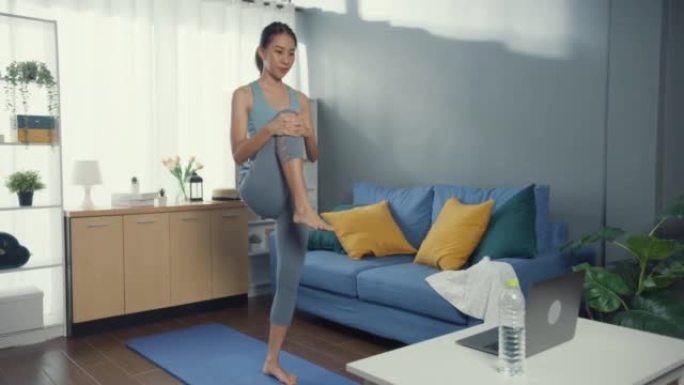 穿着运动服的年轻迷人的亚洲女性在笔记本电脑上观看健身在线视频在家里的客厅为初学者练习瑜伽。没有健身房
