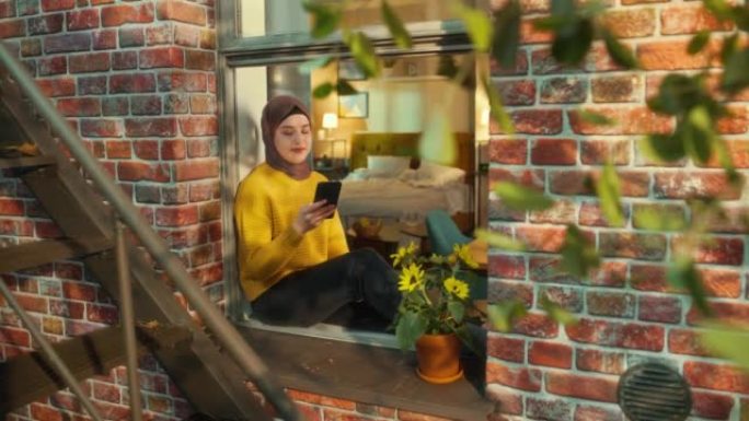 年轻的阿拉伯妇女坐在多层砖砌公寓楼的窗台上时使用智能手机。多民族女性在线与朋友联系。从房子外面向上倾