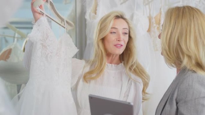 销售助理或商店老板用数字平板电脑帮助新娘商店的女人选择婚纱-慢动作拍摄