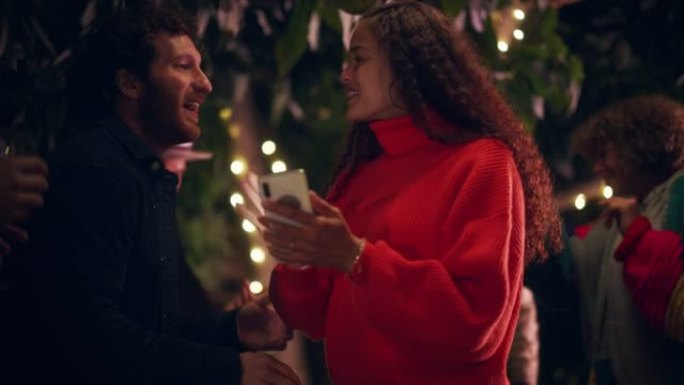 爱的混血夫妇在户外露台上使用智能手机或与朋友一起参加花园舞会。美丽的男人和女人看着电话，周末放松，玩