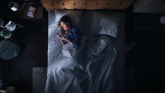 顶景卧室公寓: 美丽的年轻女子晚上在床上使用智能手机。微笑女孩浏览社交媒体，浪漫聊天，约会应用程序，