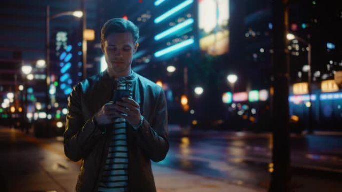 英俊的年轻人的肖像使用智能手机站在充满霓虹灯的夜城街道上。微笑的时尚金发男性使用手机进行社交媒体发布