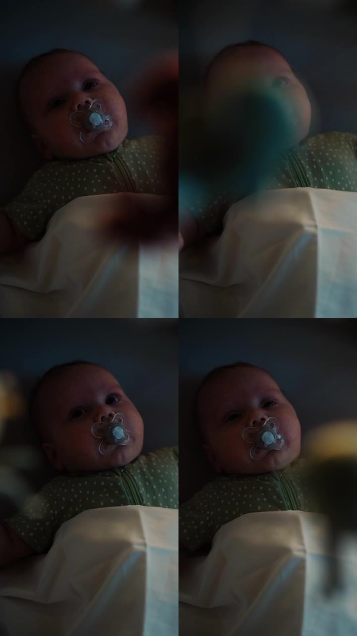 可爱的新生婴儿躺在婴儿床的背面的垂直特写肖像。顽皮兴奋的幼儿奶嘴看着旋转玩具。童年、新生活、为人父母