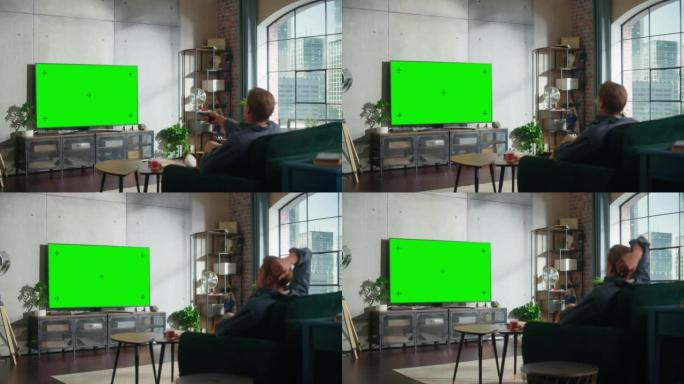 在阁楼的客厅里，一个迷人的男人坐在沙发上，打开带有控制器的绿屏模拟显示的电视。男性喜欢在家休闲。