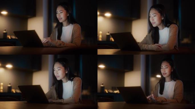 美丽地道的亚洲女人坐在舒适的厨房里的桌子旁，晚上在家使用笔记本电脑。女性微笑着浏览互联网，在社交网络
