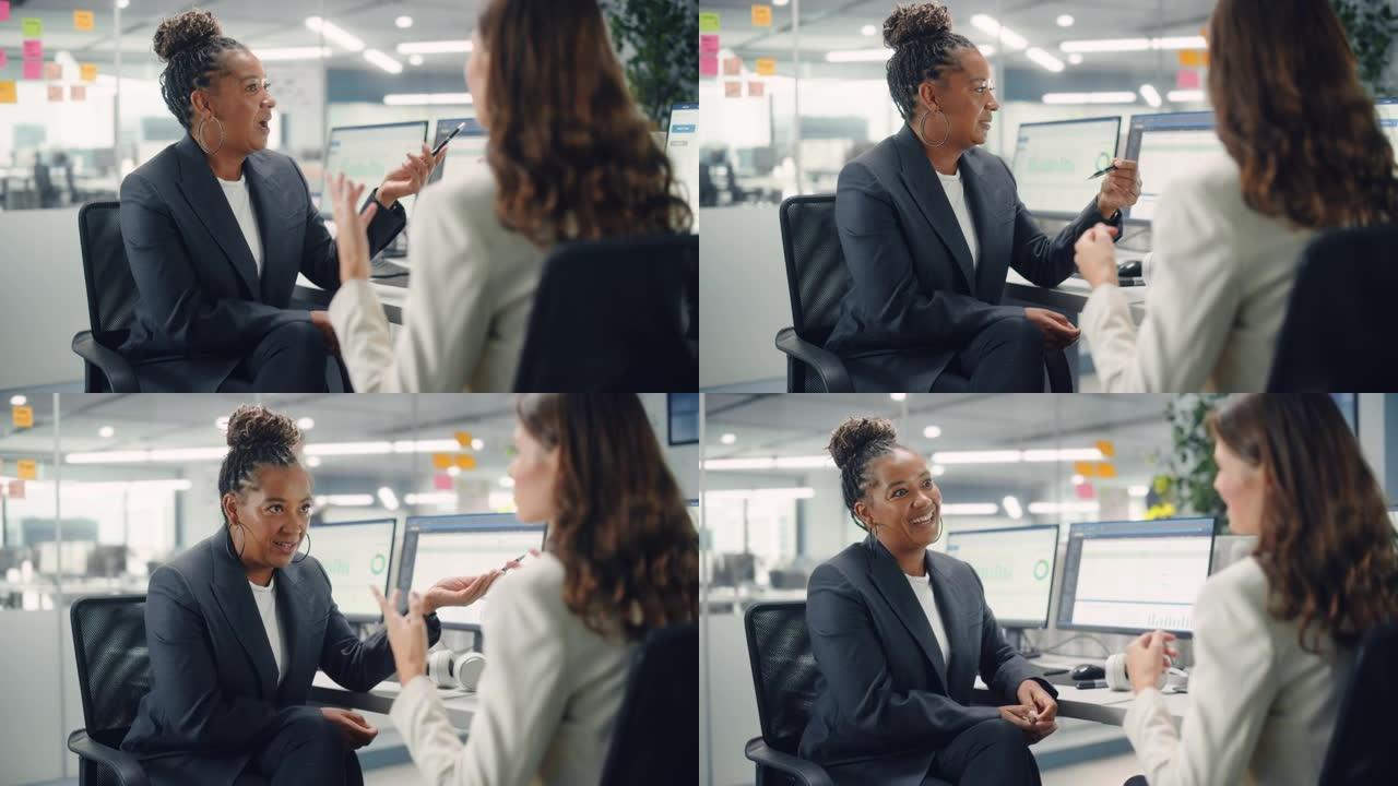 两位女同事在各种现代商务办公室的计算机上工作时，彼此亲切地交谈，笑着笑。经验丰富的经理和年轻员工讨论