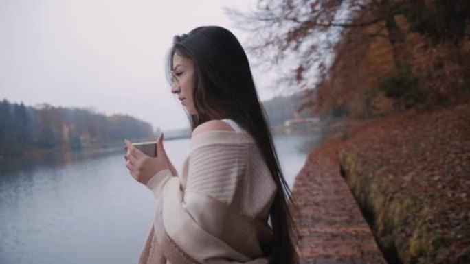 年轻女子站在湖边的木板路上，从纸杯里喝热咖啡