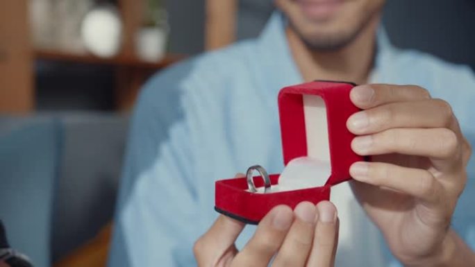 特写快乐亚洲夫妇微笑的男朋友坐在沙发上惊喜女友带着红色盒子里的订婚戒指在家里向她求婚。