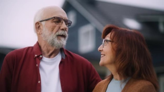 一对幸福的老年夫妇在他们的住宅区家户外摆姿势，互相拥抱的特写肖像。充满爱心的成年人看着镜头微笑，享受