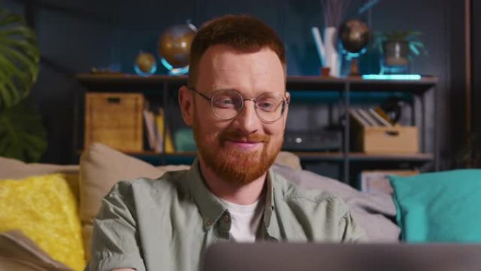 年轻快乐聪明的高加索红头发自由职业者戴着眼镜，在客厅用笔记本电脑写作。