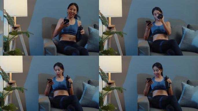 年轻的亚洲女士晚上在家客厅沙发上用手机喝纯净水。