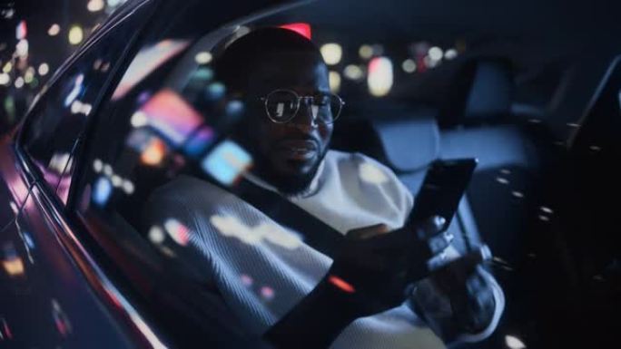 时髦的戴眼镜的黑人在晚上坐出租车的后座上下班。英俊的男性使用智能手机，在城市街道上开着霓虹灯的汽车时